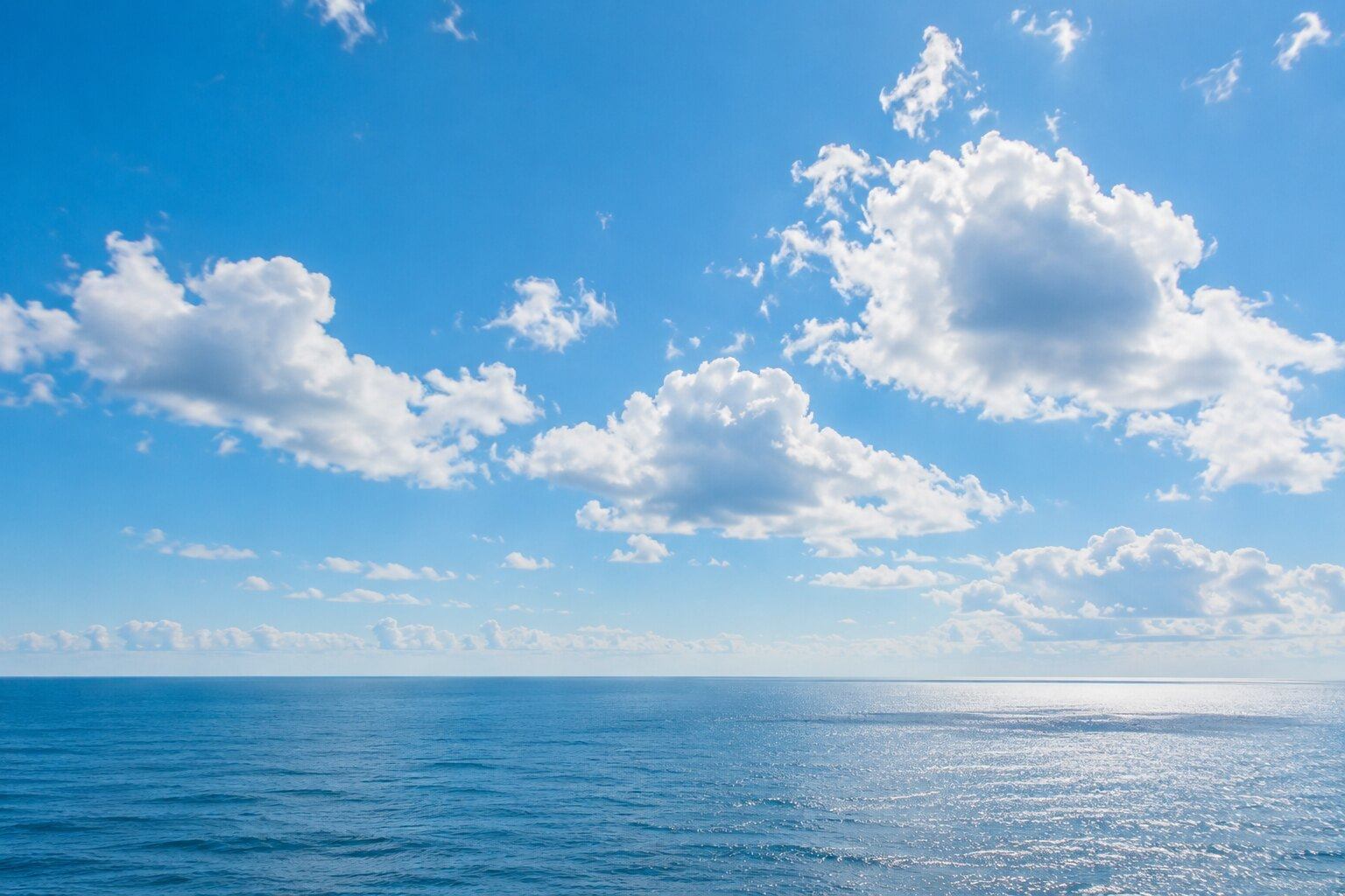 Soft white cumulus clouds in a broad light-blue sky above a thin sea horizon.